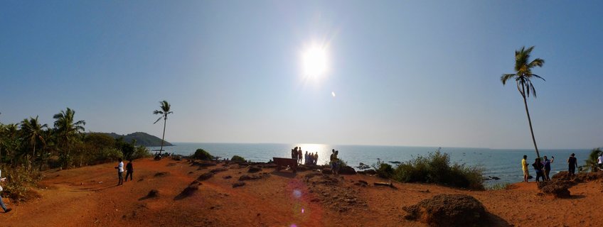 Wide coastal landscape showing people standing on red soil cliff overlooking blue ocean with palm trees and bright sun in clear sky