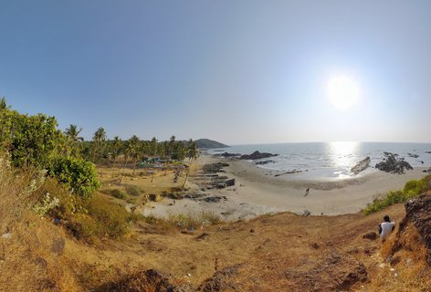 Wide coastal landscape showing a sandy beach in Goa with palm trees, rocky formations in shallow water, and a person sitting on a grassy hillside in the foreground under a clear blue sky