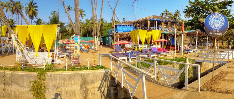 Beach restaurant in Goa with yellow fabric canopies, purple and blue lounge chairs, red umbrellas, palm trees, thatched roofs, and a wooden walkway under clear blue sky