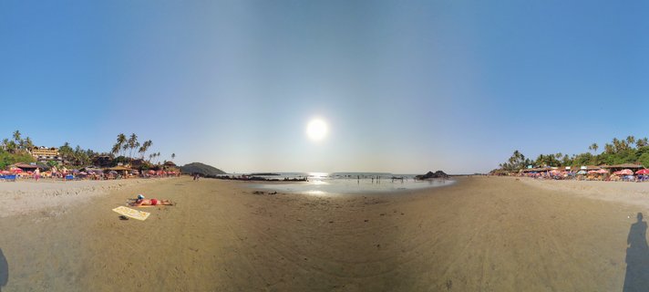 360-degree panoramic photograph of a Goa beach showing sand, ocean, palm trees, beach shacks with colorful umbrellas, and people relaxing under a clear blue sky with bright sun