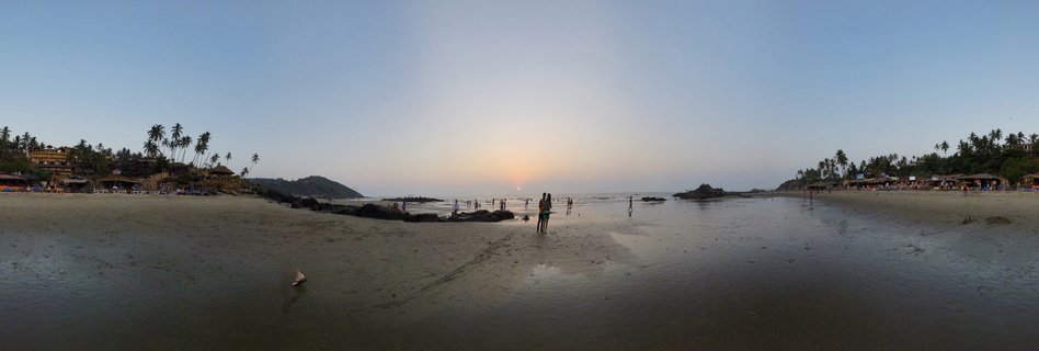 Panoramic beach scene at sunset in Goa, India showing people on wet sand, palm trees, beach shacks, and the sun setting over the ocean horizon
