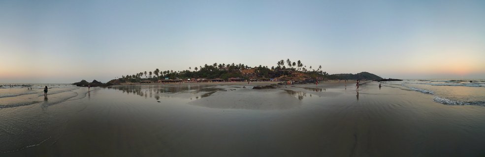 Panoramic beach scene at sunset showing wet sand with reflections, a palm tree-covered island in the center distance, people walking along the shoreline, and waves meeting the coast under a blue-to-orange gradient sky