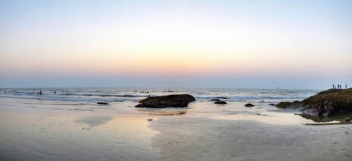 Wide beach scene at sunset in Goa showing wet sand reflections, rock formations in shallow water, ocean waves, and small figures of people in the distance under a pink and blue gradient sky