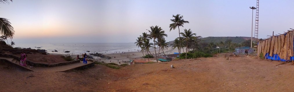 Panoramic beach scene in Goa, India showing Arabian Sea at sunset with palm trees, people on viewing platform, woman in pink sari, and traditional fishing nets drying on racks along reddish soil coastline