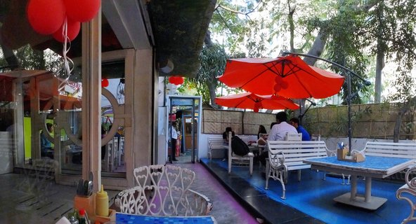 Outdoor restaurant patio with red umbrellas, white metal benches, blue floor, and patrons dining at tables near glass windows