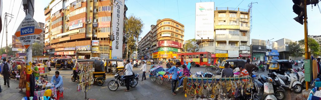 Busy urban street scene in Pune, India showing auto-rickshaws, motorcycles, street vendors with jewelry displays, pedestrians, commercial buildings with signs, and a red bus under clear afternoon sky