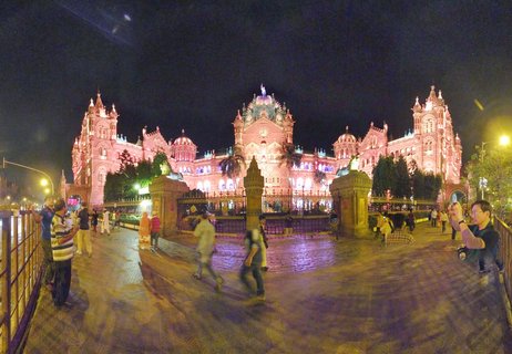 Historic railway station building illuminated in pink lights at night with people walking in the foreground courtyard