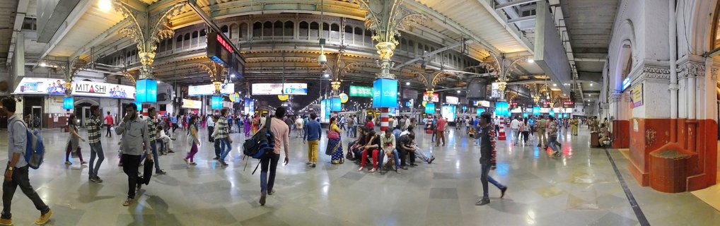 Busy interior of historic railway station in Mumbai with ornate Victorian columns, digital displays, and commuters walking across polished floors