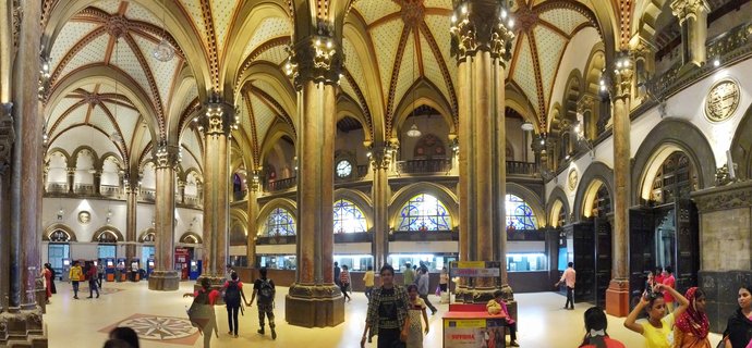 Interior view of ornate Victorian Gothic railway station with tall stone columns, vaulted ceilings with star patterns, stained glass windows, and people walking through the main hall