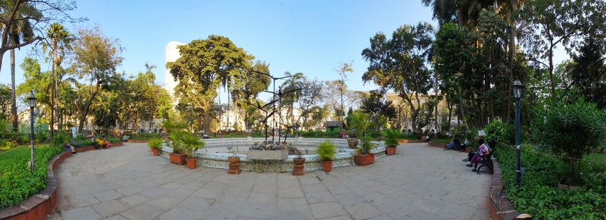Wide panoramic view of a Mumbai public park showing a circular fountain with metal sculpture at center, surrounded by stone paving, potted plants, trees, lamp posts, and people sitting on benches