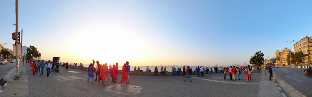 Panoramic view of people walking and sitting along Marine Drive waterfront promenade in Mumbai during sunset, with buildings and Arabian Sea visible