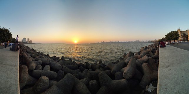 Panoramic view of sunset over the Arabian Sea from Marine Drive in Mumbai, with concrete tetrapods in the foreground, people sitting on seawalls on both sides, and the city skyline visible across the water on the horizon