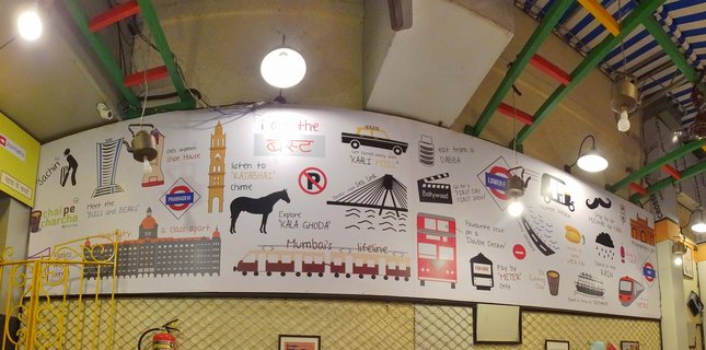 Colorful illustrated wall mural inside a cafe depicting Mumbai landmarks including Gateway of India, local train, double-decker bus, auto-rickshaw, Sea Link bridge, and text references to neighborhoods and local culture