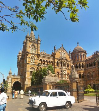 Victorian Gothic railway station building with ornate stone towers, domes and arched windows, white classic car parked in front, people walking on paved courtyard, clear blue sky above