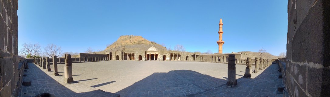 Stone courtyard with rows of pillars leading to arched mosque building with tall red minaret and hill in background under blue sky