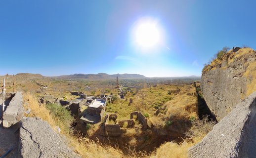 Panoramic photograph showing ancient stone fort ruins with weathered walls, a tall minaret tower in the distance, dry golden grassland, rocky hills, and bright sun against clear blue sky in Aurangabad, India.