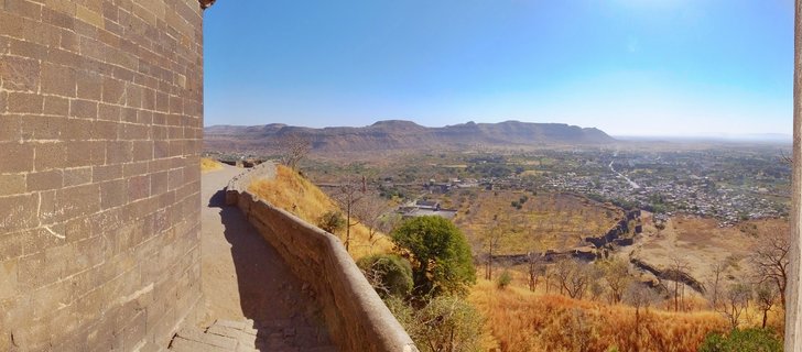 Stone fortification wall on the left frames a panoramic view of Aurangabad city in the valley below, with dry golden grasslands, scattered trees, and distant mountains under clear blue sky