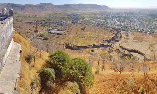 Elevated view showing medieval stone fort walls extending across dry grassland with a tall red minaret tower, distant town, and hills on the horizon under clear blue sky in Aurangabad, India