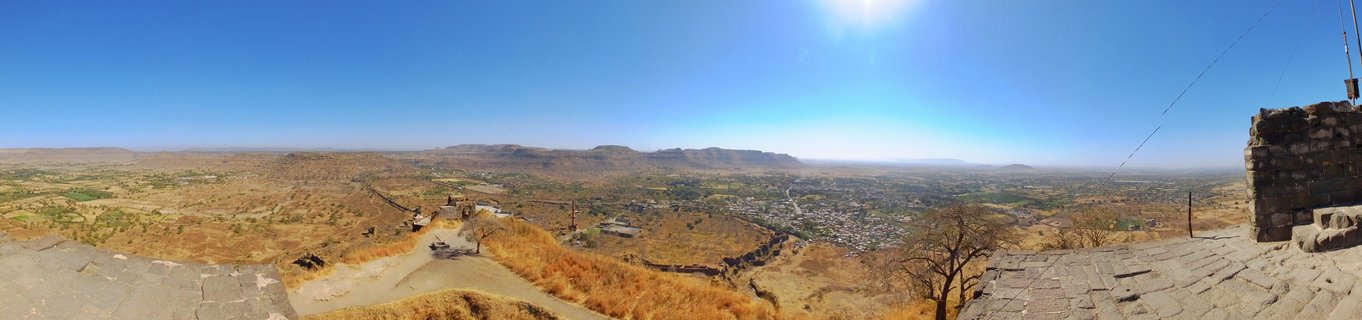 Wide panoramic landscape showing stone fortress walls overlooking a dry valley with distant mountains and a small town under clear blue sky in Aurangabad, India