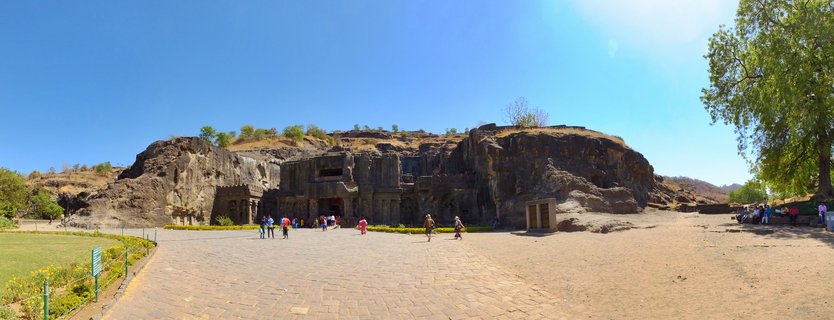 Wide view of the Kailasa Temple carved into a massive rock cliff face at Ellora Caves, with tourists walking on stone pavement in the foreground under a clear blue sky
