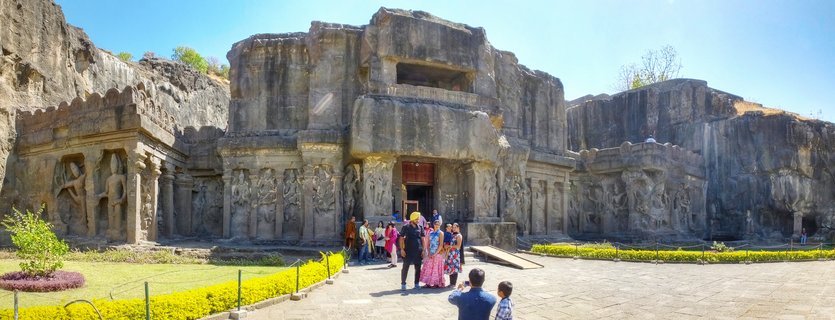 Wide view of the Kailasa Temple at Ellora Caves showing the massive rock-cut stone structure carved into a cliff face with visitors in colorful clothing walking on the courtyard in front under a clear blue sky