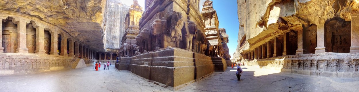 Panoramic view of the Kailasa Temple at Ellora Caves showing the monolithic rock-cut structure with elephant sculptures, columned galleries, and visitors walking through the courtyard under bright daylight