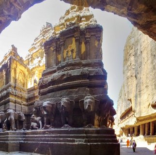 View through cave opening of Kailasa Temple showing carved elephant sculptures at base with temple towers rising behind, tourists visible in background courtyard