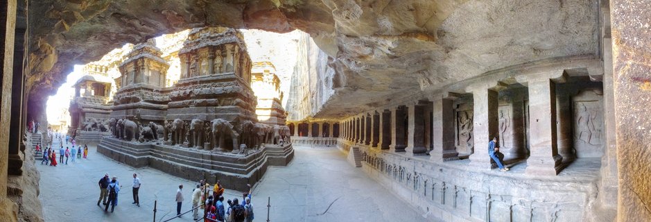 Panoramic view inside Kailasa Temple cave showing the monolithic stone temple structure with carved elephant sculptures at its base, surrounded by rock-cut colonnades and visitors walking through the courtyard