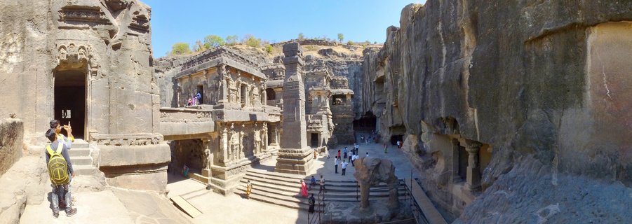 Rock-cut temple complex carved into cliff face with tourists walking on stone steps and courtyards under clear blue sky at Ellora Caves