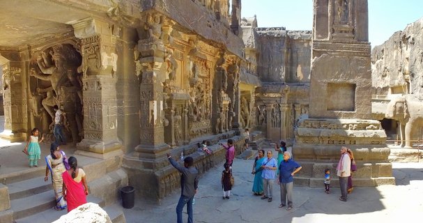 Tourists walking among ancient carved stone pillars and temple walls at Ellora Caves in India, featuring intricate Hindu sculptures and rock-cut architecture under bright sunlight