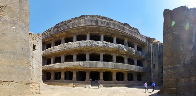 Panoramic exterior view of Tin Tal cave at Ellora Caves showing three-story rock-cut Buddhist monastery facade carved into basalt cliff with stone columns and balconies, tourists visiting under clear blue sky in Aurangabad, Maharashtra, India