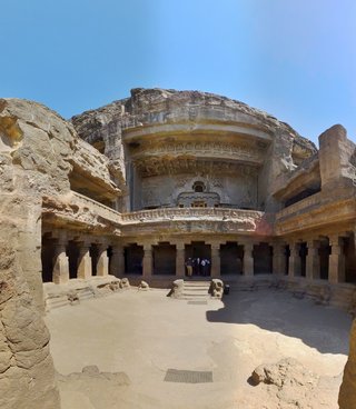 Ancient rock-cut Buddhist temple carved into cliff face with two-story pillared courtyard, stone columns, carved sculptures, and visitors at entrance under clear blue sky