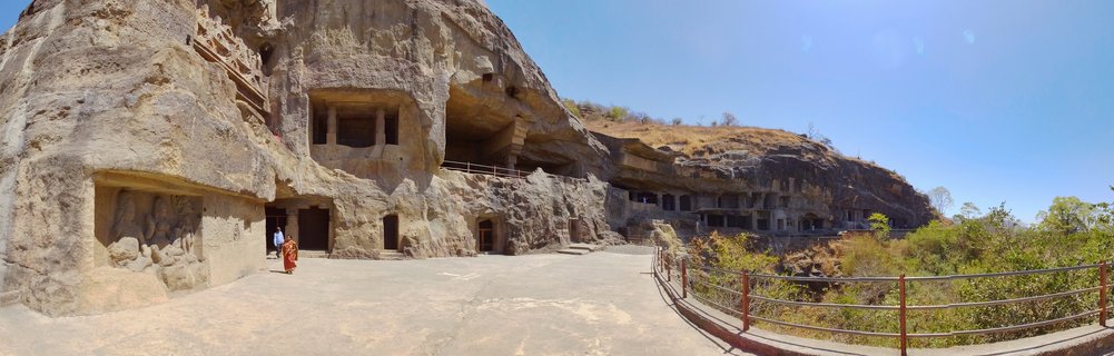 Wide view of ancient cave temples carved into rocky cliff face with stone pillars, arched entrances, paved terrace walkway, and metal safety railings under clear blue sky in Aurangabad, India.