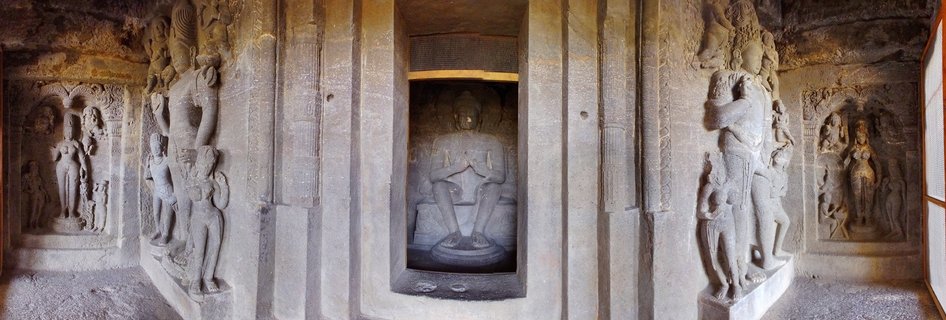Interior of ancient Buddhist cave temple showing central seated Buddha statue in stone shrine, flanked by carved pillars and wall relief sculptures of deities and figures