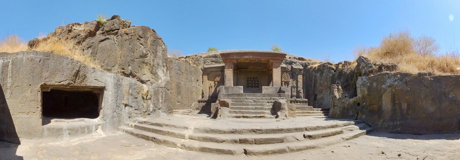 Ancient rock-cut Buddhist caves carved into brown volcanic stone cliff with stone steps, pillars, and cave entrances under clear blue sky in Aurangabad, India