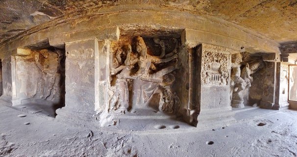 Interior of ancient rock-cut cave temple showing carved stone pillars with Hindu deity sculptures, natural rock ceiling, and stone floor with circular indentations