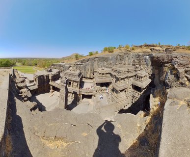Aerial view of the Kailasa Temple at Ellora Caves showing the multi-level rock-cut Hindu temple complex carved from basalt cliff with tourists visible on the stone courtyards under clear blue sky