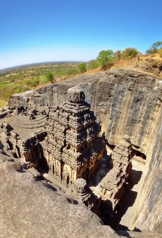 Aerial view of the Kailasa Temple carved from rock at Ellora Caves with surrounding cliffs and distant green landscape under clear blue sky