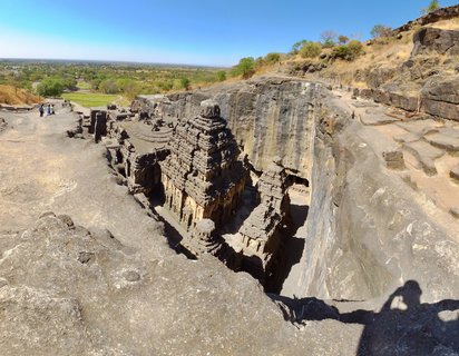 Aerial view of the Kailasa Temple carved from dark volcanic rock at Ellora Caves, with tourists walking on surrounding stone pathways under clear blue sky