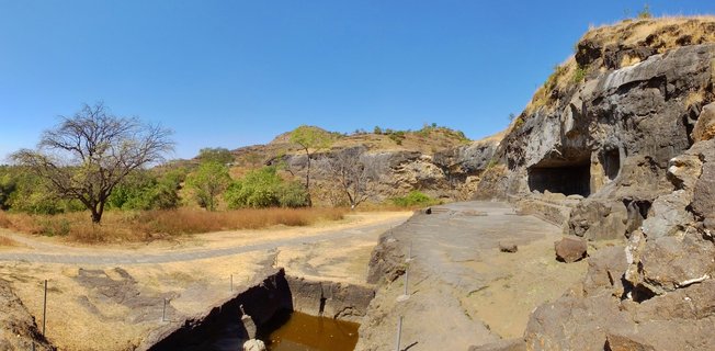 Ancient rock-cut Buddhist caves carved into rocky hillside with stone pathways, water cistern, sparse vegetation, and clear blue sky in Aurangabad, India