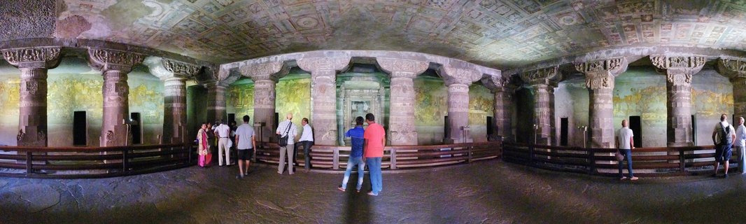 Interior view of ancient rock-cut cave temple with carved stone pillars, painted ceiling frescoes, and tourists observing the historic Buddhist artwork