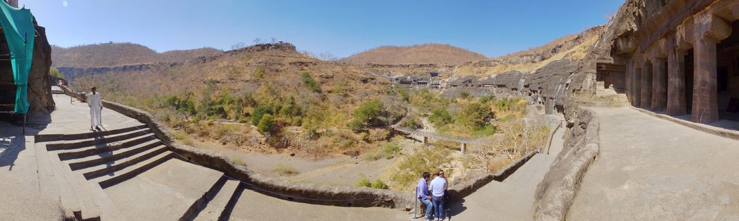 Panoramic view of Ajanta Caves showing ancient rock-cut Buddhist monuments carved into volcanic cliff with tourists walking on pathways, dry hills and blue sky in Aurangabad, India