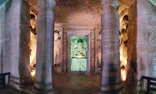 Interior view of Ajanta Caves showing a large seated Buddha statue in the center with hands in teaching gesture, flanked by carved stone pillars. Multiple smaller Buddha sculptures are visible on the cave walls. Warm artificial lighting illuminates the ancient rock-cut Buddhist temple interior.