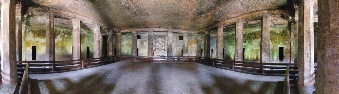Panoramic interior view of ancient rock-cut Buddhist cave temple with carved stone pillars, weathered wall murals, wooden railings, and Buddha statue at far end