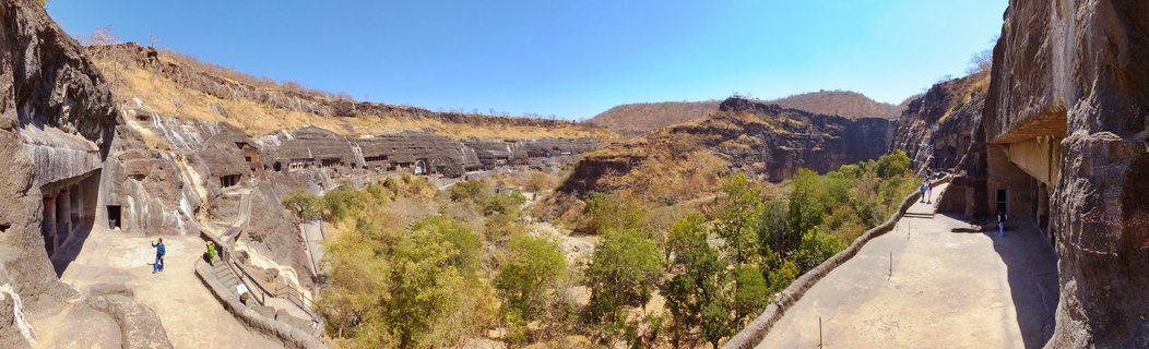 Wide panoramic photograph showing the Ajanta Caves carved into brown rock cliffs forming a horseshoe-shaped valley with green trees below, walkways with visitors, and clear blue sky above