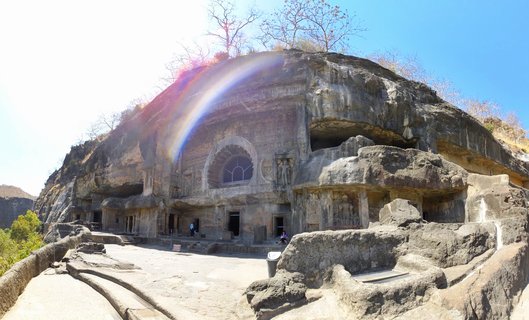 Ancient Buddhist cave temples carved into a rocky cliff face with arched windows, stone pillars, and visitors walking on a stone pathway under a clear blue sky
