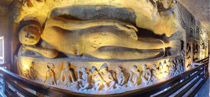 Large reclining Buddha sculpture carved into cave wall with warm lighting, surrounded by smaller seated disciple figures, wooden railing in foreground at Ajanta Caves