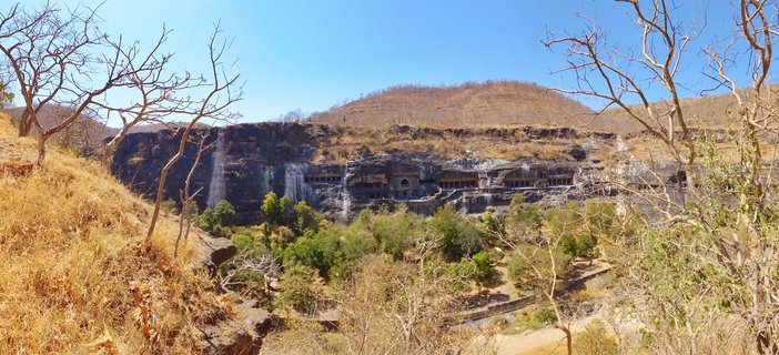 Wide view of ancient rock-cut cave temples carved into cliff face with multiple columned openings, surrounded by dry grassy hills and sparse trees under blue sky in Aurangabad India