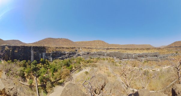 Panoramic view of Ellora Caves showing ancient rock-cut temple structures carved into a dark basalt cliff face with dry hills and sparse vegetation in the foreground under clear blue sky
