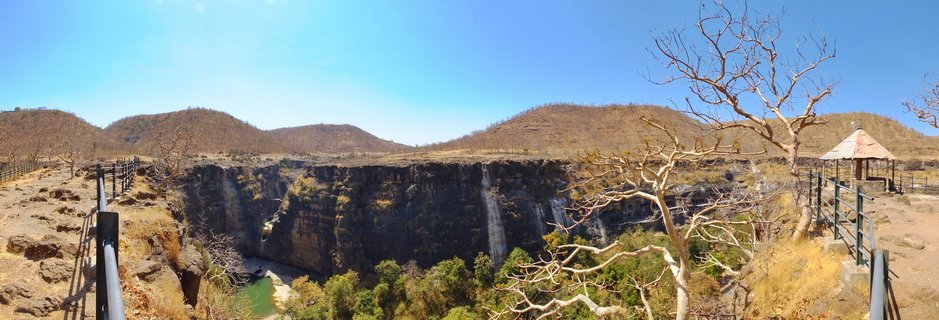 Wide panoramic landscape showing a deep gorge with waterfalls flowing down dark rock cliffs, surrounded by dry brown hills, metal railings and a gazebo in the foreground, clear blue sky above